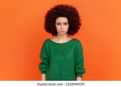 Sad unhappy woman with Afro hairstyle wearing green casual style sweater looking at camera with pout lips, being upset of bad news. Indoor studio shot isolated on orange background.