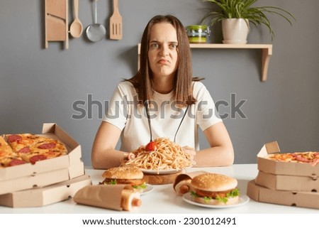 Sad unhappy woman wearing white t- shirt siting at table with fast food in kitchen looking at camera with displeased face felling overeating unhealthy nutrition.