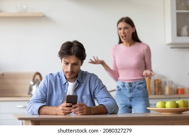 Sad Unhappy Young Caucasian Husband With Smartphone Ignoring Upset Angry Scolding Wife In Minimalist Kitchen Interior. Jealousy, Device Addiction, Relationship Problems And Quarrel At Home, Free Space