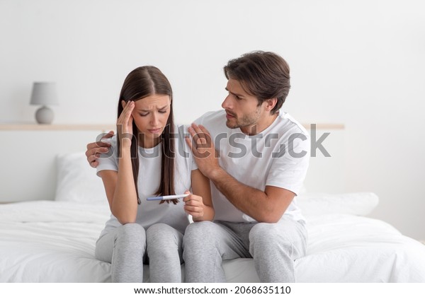 Sad unhappy young caucasian husband calm his wife\
on bed, lady look at pregnancy test in bedroom interior, free\
space. Infertility, unwanted pregnancy, health problems, family\
planning and support