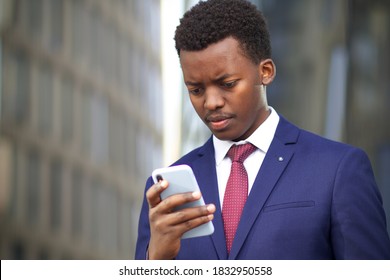 Sad Upset Frustrated Black African Afro American Man Looking At Screen Of His Smartphone. Unhappy Office Worker, Businessman In Formal Suit Outdoors. Problems With Cell Mobile Phone. Negative News. 