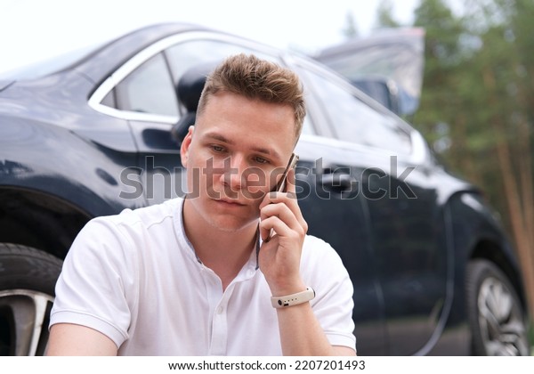 Sad\
upset guy driver sitiing near broken car after road accident,\
crash, calling emergency on cell mobile phone talking on\
smartphone. Young desperate man got into\
accident