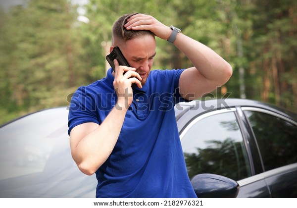 Sad upset guy driver is standing near broken car\
after road accident, crash, calling emergency on cell mobile phone\
talking on smartphone. Young desperate man got into accident\
holding head with hand 