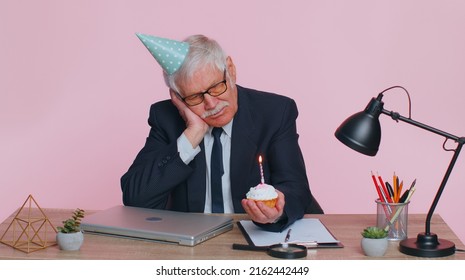 Sad Upset Lonely Senior Businessman In Festive Hat Celebrating Birthday Party Alone, Feel Frustrated Despair Disappointed Sadness Holds Cake With Candle At Office. Elderly Man Isolated On Pink Studio