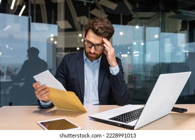 Sad and upset mature businessman received an envelope by mail with a bad news notification letter, boss in business suit working inside office using laptop wearing glasses and beard. - Powered by Shutterstock