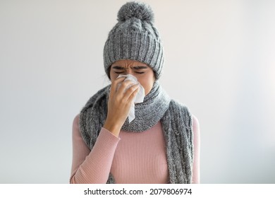 Sad Upset Millennial Indian Female In Sweater, Hat And Scarf Blows Nose In Napkin On Gray Wall Background, Free Space. Illness, Flu, Cold, Covid-19 Virus And Health Problems In Winter Due Quarantine