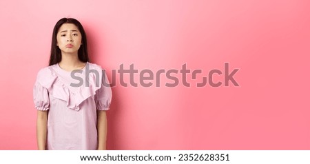Sad whining asian girl pouting and frowning, looking upset about something unfair, complaining, standing in dress against pink background.