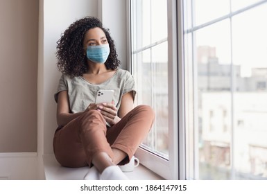 Sad woman alone during coronavirus pandemic wearing face mask indoors at home for social distancing. Mixed race girl looking at window. Anxiety, stress, lockdown, mental health crisis concept