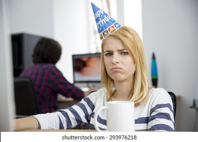 Sad Woman With Birthday Hat Sitting At The Table In Office And Looking At Camera