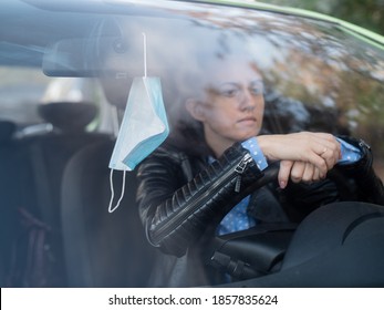 Sad woman driver sits in the car and holds his hands on the steering wheel of the car. Medical mask hanging on the mirror in the car