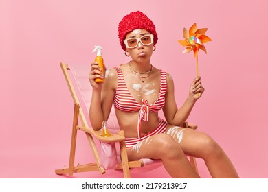 Sad Woman With Fit Body Has Bad Mood Holds Suncreen Lotion And Windmill Gets Suntan At Beach Dressed In Swimsuit Sits On Deck Chair Isolated Over Pink Background Spends Summer Vacation At Seaside