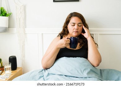 Sad woman in her 20s with obesity suffering from a headache or migraine while drinking hot tea in bed