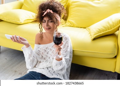 Sad Woman Holding Glass Of Wine And Smartphone While Crying In Living Room