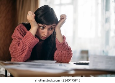 Sad woman holding her head over utility bills. The concept of rising prices for heating, gas, electricity. A young worried dissatisfied girl looks at papers does not expect a high cost of services - Shutterstock ID 2140475945