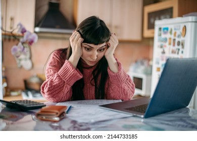 Sad woman holding her head over utility bills in front of a laptop. The concept of rising prices for heating. A young worried girl looks at papers, documents, does not expect a high cost of services. - Shutterstock ID 2144733185