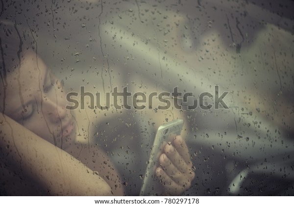 Sad woman looking in the mobile phone and reading\
massega in the window with rain drop in the car. Face of young\
female behind rain car window. Loneliness and depression concept.\
Psychology 