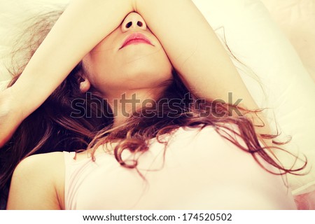 Sad woman is lying in bed with her arm on head and eyes. Young woman with long hair, wears pink underwear.