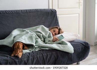 Sad Woman Lying On Sofa With Dog, Using Smart Phone In Hands Wait For Boyfriend Call Or Text, Feels Depressed. Thoughtful Female Think About Relationships Problems. Mental Health, Loneliness Concept.