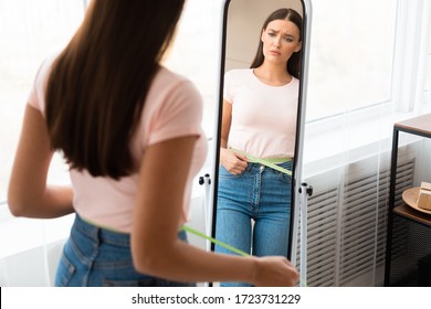 Sad Woman Measuring Waist With Tape After Weight Gain Standing In Front Of Mirror At Home. Slimming Concept. Free Space - Shutterstock ID 1723731229