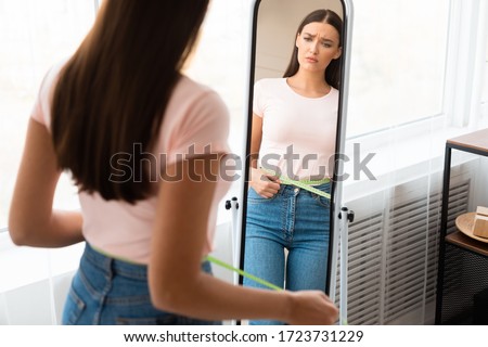 Sad Woman Measuring Waist With Tape After Weight Gain Standing In Front Of Mirror At Home. Slimming Concept. Free Space