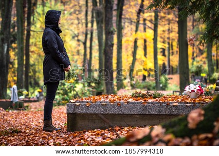 Sad woman is mourning for dead person at grave in cemetery. Lonely mourner in autumn graveyard.  Grief and cry for dead people