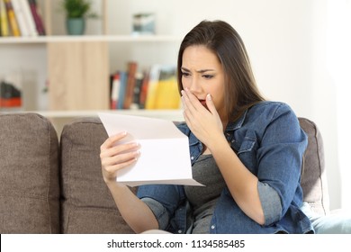 Sad woman reading bad news in a paper letter sitting on a couch in the living room at home