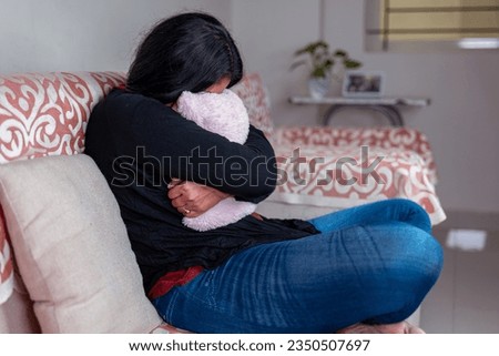 A sad woman sits on a couch, tightly hugging a pillow. She looks lost and alone