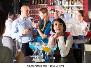 Sad Woman Sitting Alone At Office Party In Nightclub In Background With Cheerful Workmates