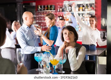 Sad Woman Sitting Alone At Office Party In Nightclub In Background With Cheerful Workmates