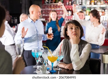 Sad Woman Sitting Alone At Office Party In Nightclub In Background With Cheerful Workmates