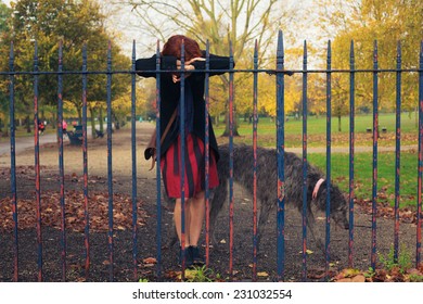 A Sad Woman Is Standing By A Fence In The Park With A Dog