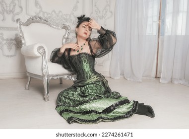 Sad woman wearing green medieval vintage Victorian Style dress sitting on the floor near armchair - Shutterstock ID 2274261471