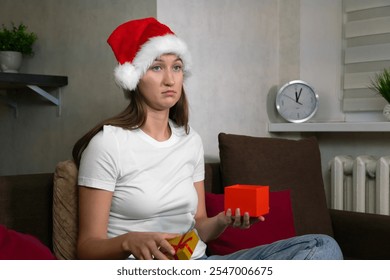 Sad woman wearing santa hat opening christmas gift box at home, expressing disappointment with the present she received - Powered by Shutterstock