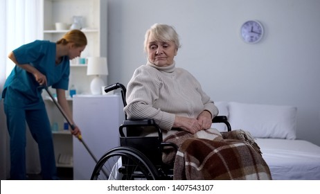 Sad Woman In Wheelchair Watching Hospital Janitor Cleaning Room, Medical Center