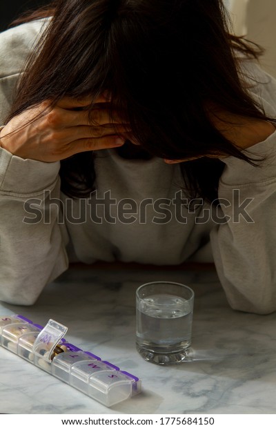 A sad women with her head down and hands on her head is\
seen right before she takes her daily medication for chronic\
disease. She stares at a glass of water and the pill box next to\
it. 
