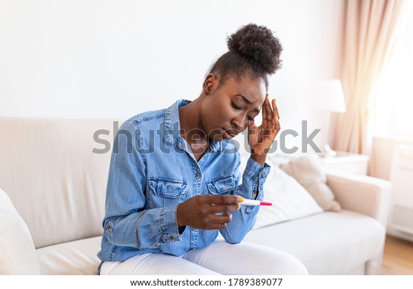 Sad, worried african\
american woman checking her recent pregnancy test, sitting on couch\
at home. Maternity, child birth and family problems concept.\
unwanted pregnancy