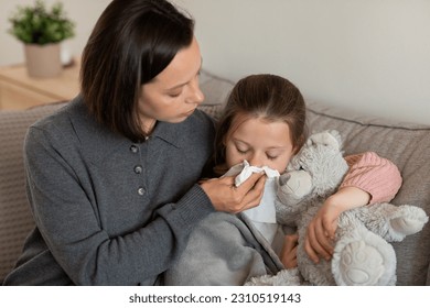 Sad worried caucasian mature mother blows nose to napkin to little daughter, lies on sofa in living room interior. Health care, treatment and medicine at home, parent support