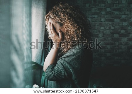Sad and worried woman side portrait at home in the dark looking outside the window. Worried and lonely female alone indoor. Concept of end of relationship and unsuccessful emotion. Lady touching face