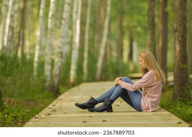 Sad young adult woman sitting alone on wooden trail at birch tree forest in autumn day. Thinking about life problem at nature park. Suffering from relationship break up. Side view.
