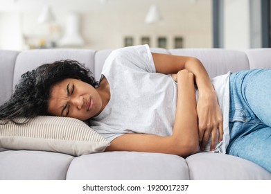 Sad Young African American Woman Wearing Casual Clothes Suffering From Menstrual Pain, Feeling Sick To Her Stomach, Holding Belly, Having Abdominal Cramps During Period And Lying Down On Bed At Home