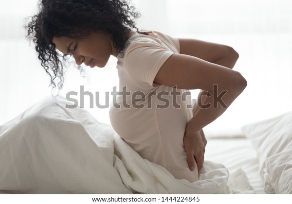 Sad young african woman touching back feeling\
backache morning discomfort low lumbar muscular kidney pain sit on\
bed after bad sleep waking up on uncomfortable mattress bending\
from backpain concept