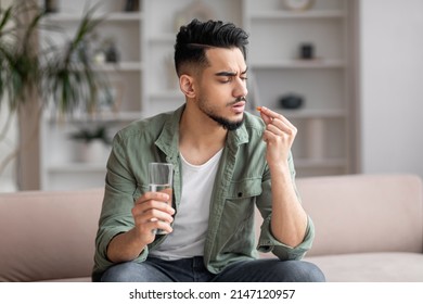 Sad young arab man with beard in casual sits on sofa with glass of water and looks at pill in hand in living room interior. Health care, disease treatment and painkiller at home, covid-19 outbreak - Shutterstock ID 2147120957