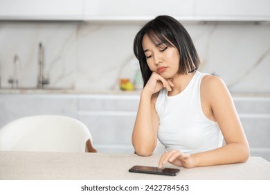 Sad young Asian girl sitting thoughtfully at the kitchen table with smartphone in hand - Powered by Shutterstock