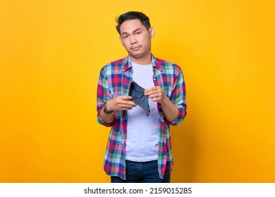 Sad young Asian man in plaid shirt showing his empty wallet and looking at camera isolated on yellow background