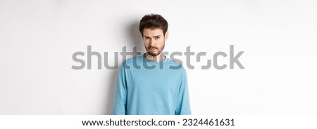 Sad young bearded man sulking, looking upset at camera, offended by someone, standing against white background.