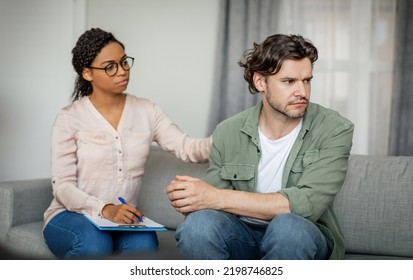 Sad Young Black Lady Doctor Calms Unhappy Despaired Caucasian Male Patient In Office Clinic Interior. Medical Support, Psychological Therapy, Session With Professional, Mental Help For Suffer Client