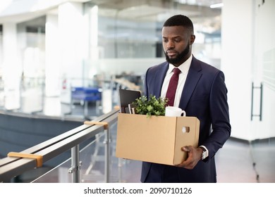 Sad young black man got fired, walking by office building and holding box with his belongings, copy space. Terminated african american guy in suit lost his job, leaving workplace, copy space