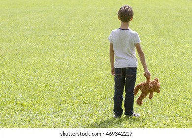 Sad Young Boy Is Holding A Brown Teddy Bear And Standing On The Meadow. Back View. Copy Space. Sadness, Fear, Frustration, Loneliness Concept