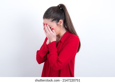Sad young caucasian woman wearing red shirt over white background crying covering her face with her hands.
