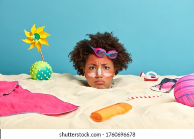 Sad young female model covered with sand, only head is visible, applies sunscreen on sunburn skin, worried because of being completely sunburned, poses at seaside, rests with beach items around